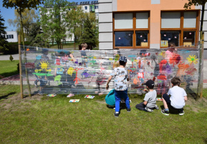 Dzieci malują na foli, w tle drzewa oraz pomarańczowy budynek przedszkola